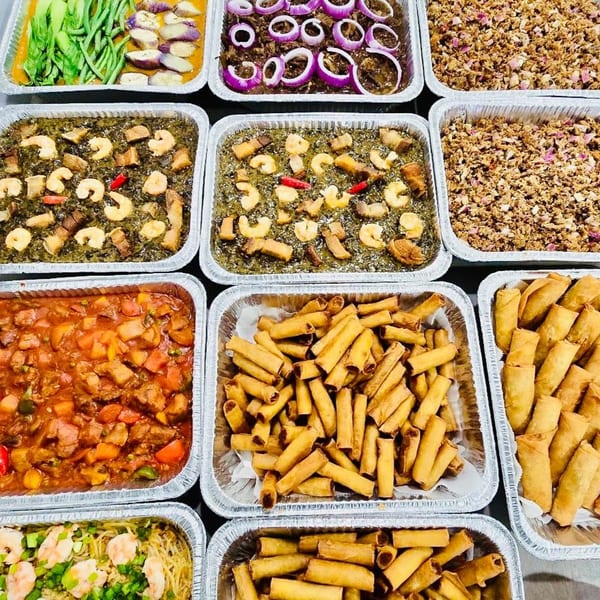 A catering spread of various Filipino dishes in foil trays, including lumpia, sisig, and vegetable stews.