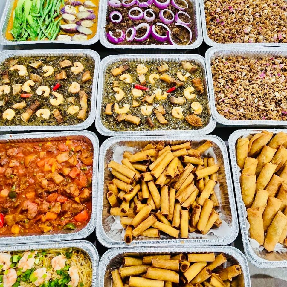 A catering spread of various Filipino dishes in foil trays, including lumpia, sisig, and vegetable stews.